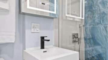 Une salle de bain moderne avec une douche à l’italienne en verre, un lavabo blanc doté d’un robinet noir et un pare-baignoire en marbre.