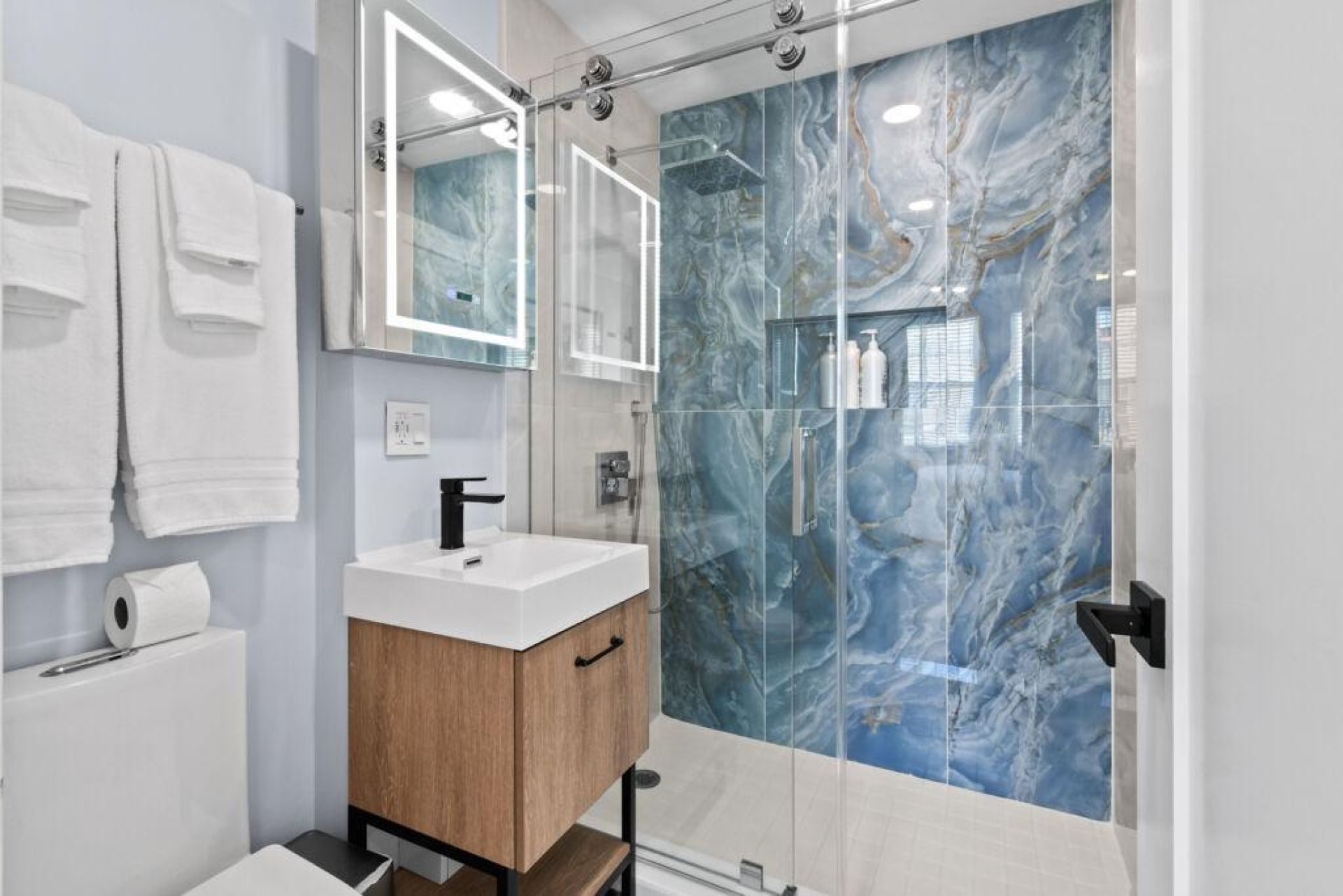 A modern bathroom with a marble shower wall, a white sink, and a wooden vanity.