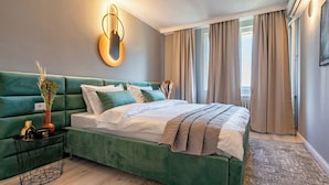 A modern bedroom with a green upholstered headboard, a neatly made bed with white and grey bedding, a small side table with a vase and a lamp, and a large window with curtains. - Golden Glow Hideaway (Bucharest)