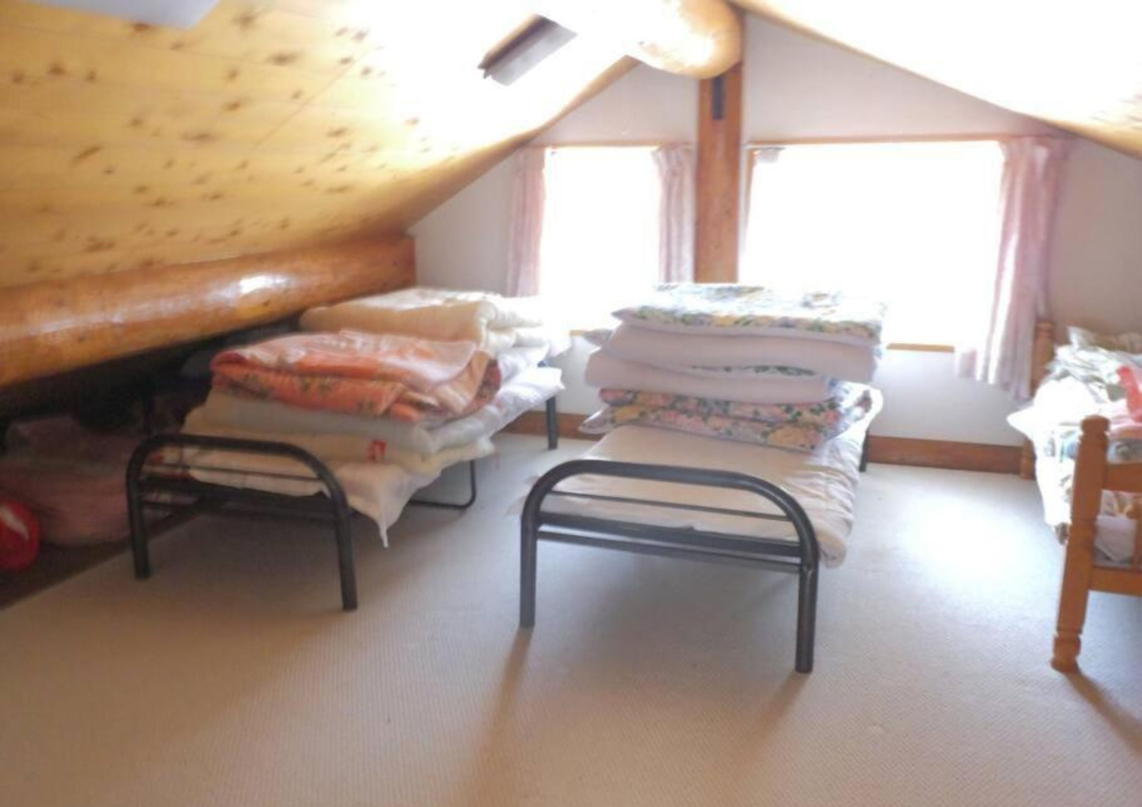 Une petite pièce agréable, avec un plafond en pente, un lit recouvert d’une couette à motifs floraux, une table de chevet et une fenêtre avec des rideaux.
