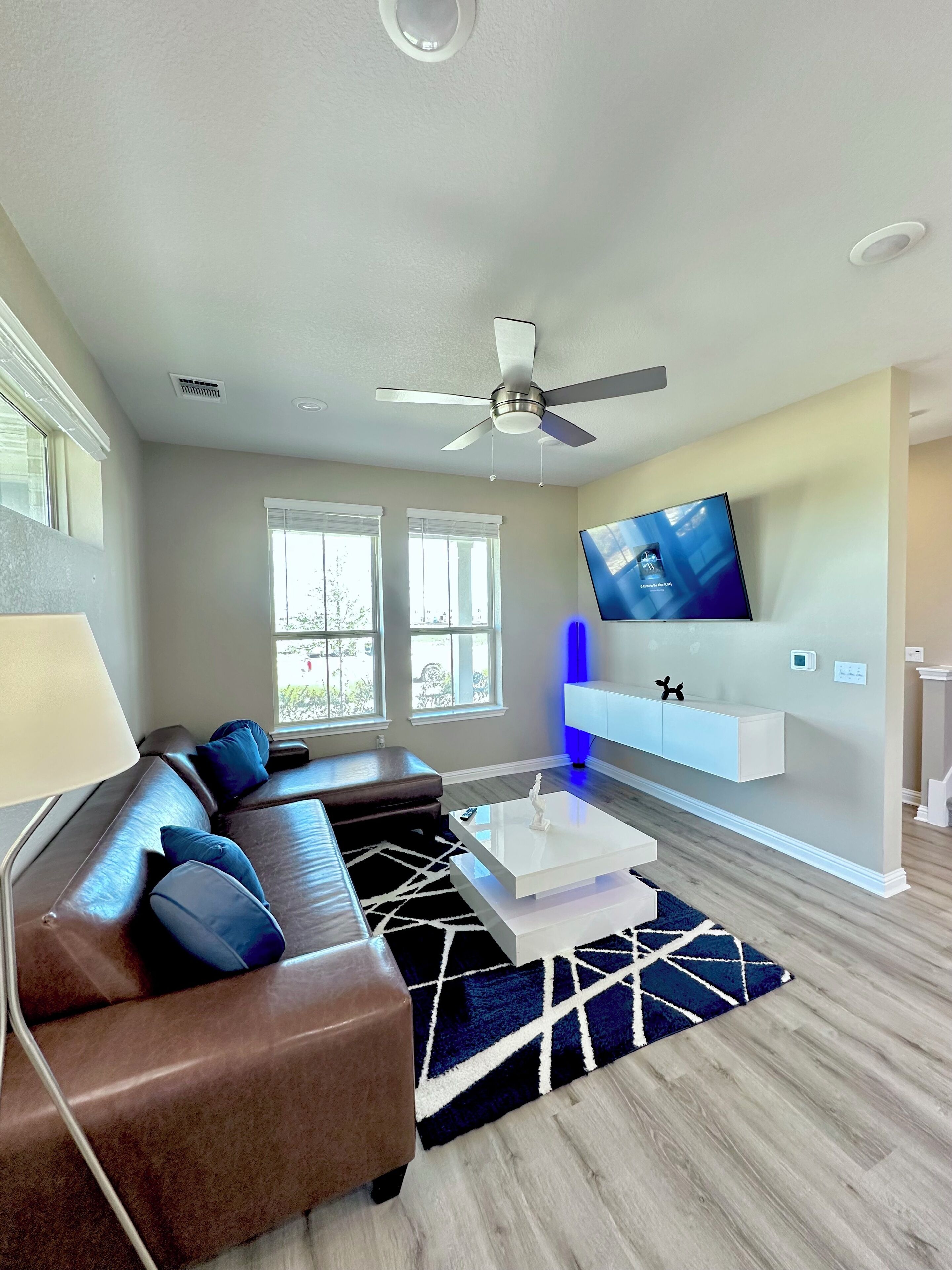 Un salon moderne avec un canapé d’angle, un téléviseur à écran plat fixé au mur, un ventilateur de plafond et un tapis à motifs.
