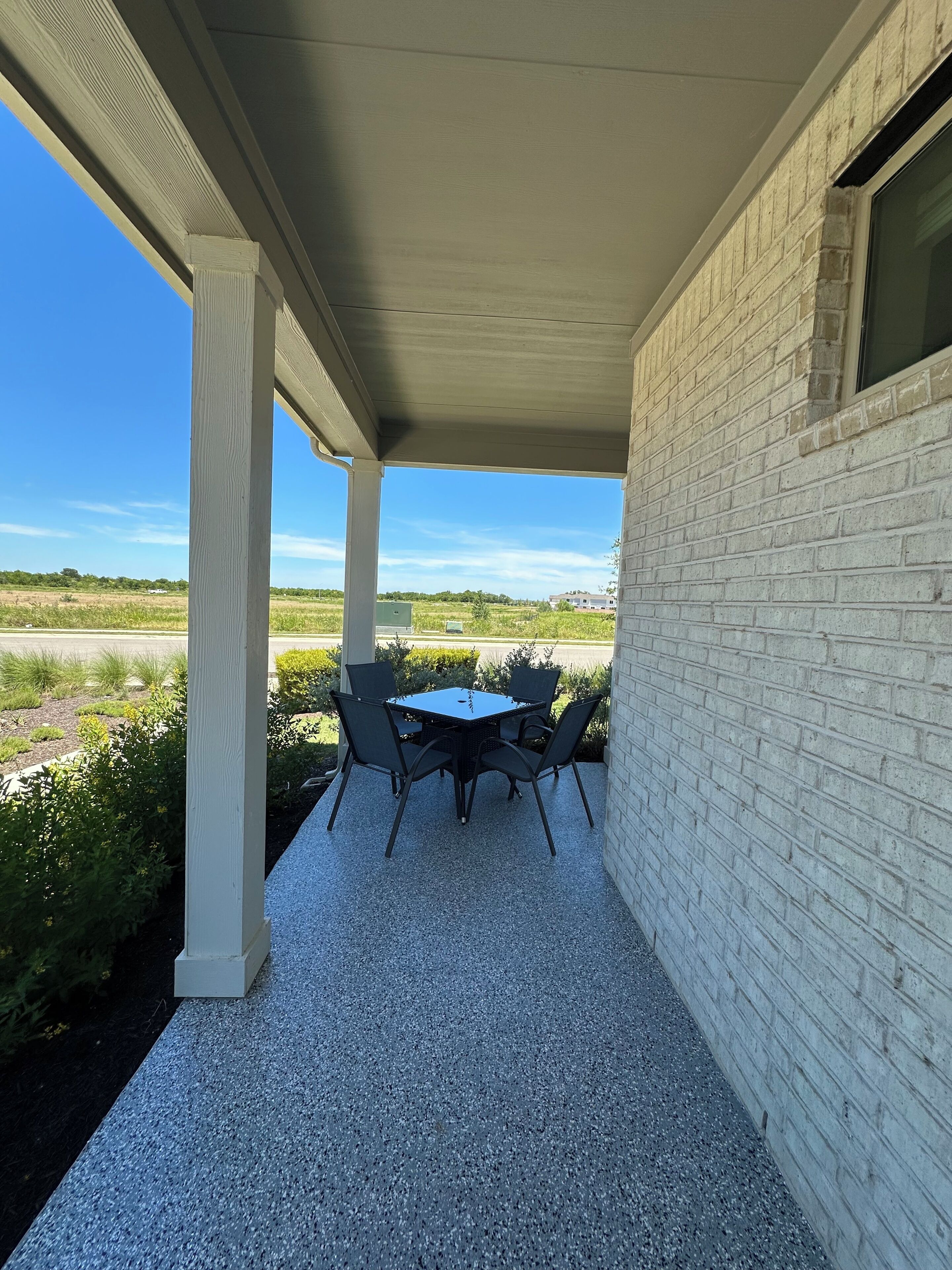 Ein überdachter Patio-Bereich mit Tisch und Stühlen, eine weiße Backsteinmauer und Blick auf ein Feld.