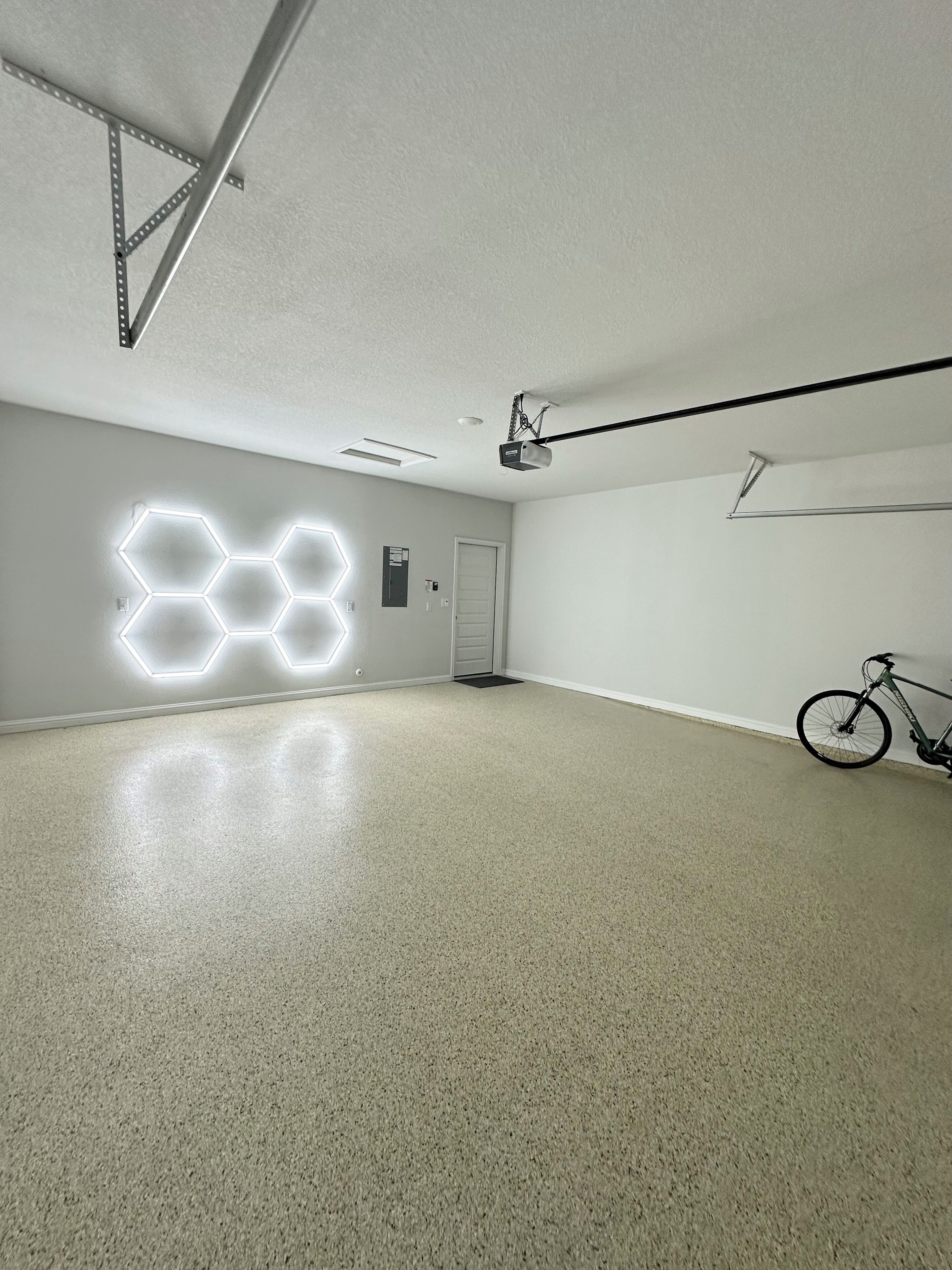 A spacious, well-lit room with a hexagonal light installation on the wall, a ceiling-mounted projector, and a bicycle leaning against the wall.