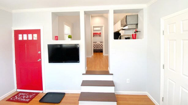 Un pasillo con una puerta roja, un televisor de pantalla plana montado en la pared, una escalera y una cocina visible a través de una puerta abierta.