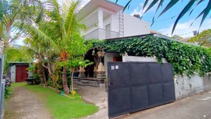 Propiedad residencial con entrada protegida por un muro, camino de entrada pavimentado y abundante vegetación, incluyendo palmeras y un muro cubierto de hiedra.