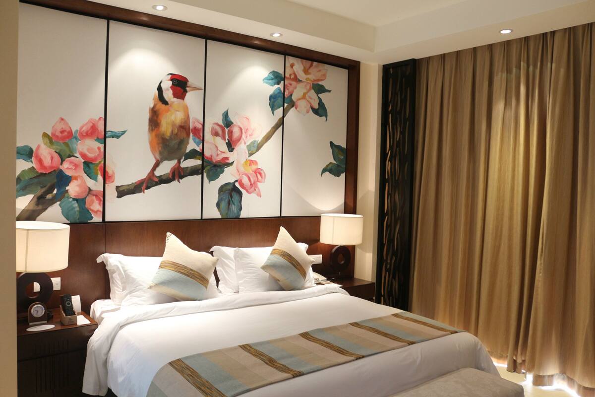 Una habitación de hotel con una cama grande, dos mesitas de noche, una lámpara de noche, un cuadro de pájaros y flores en la pared y una cortina.