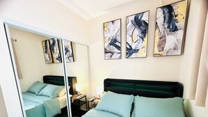 A hotel room with a mirrored closet, a bed with light blue bedding, two bedside tables with lamps, and abstract artwork on the walls. - Lake Murray 3BR Retreat with AC and Skylight (La Mesa)
