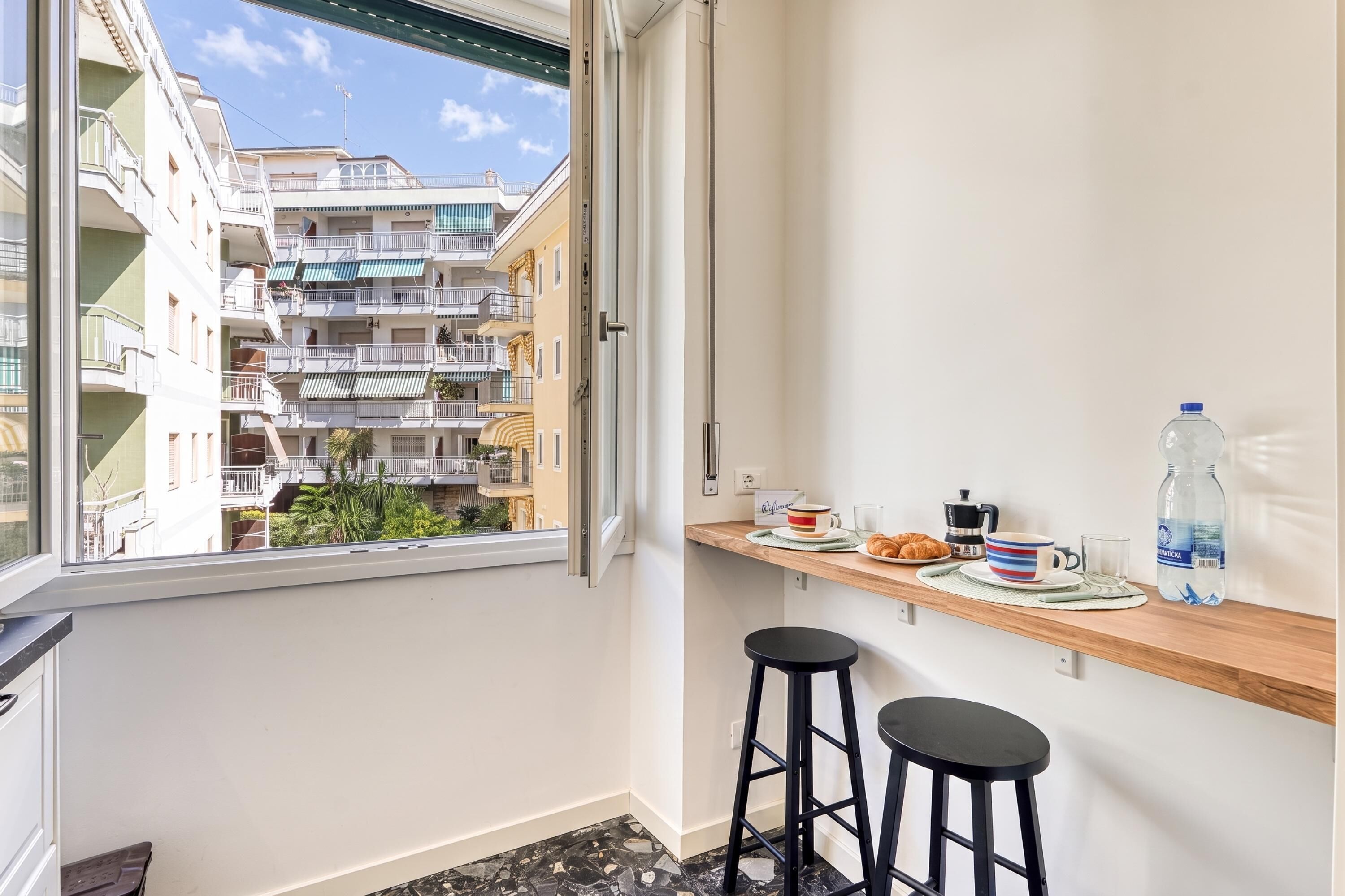 A balcony with a view of a multi-story building, a wooden shelf with breakfast items, and a black stool.