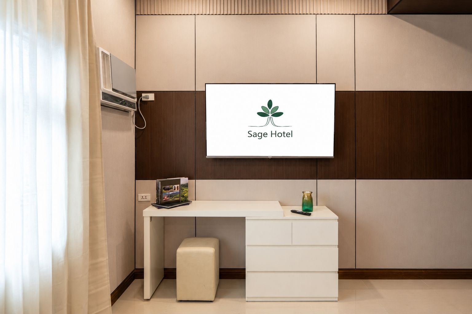 Et hotelreceptionsområde med et fladskærms-tv, der viser hotellets logo, et hvidt skrivebord med skuffer, en lille skammel og en fjernbetjening.