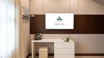 Un'area reception di un hotel con una TV a schermo piatto che mostra il logo dell'hotel, una scrivania bianca con cassetti, un poggiapiedi e un telecomando.
