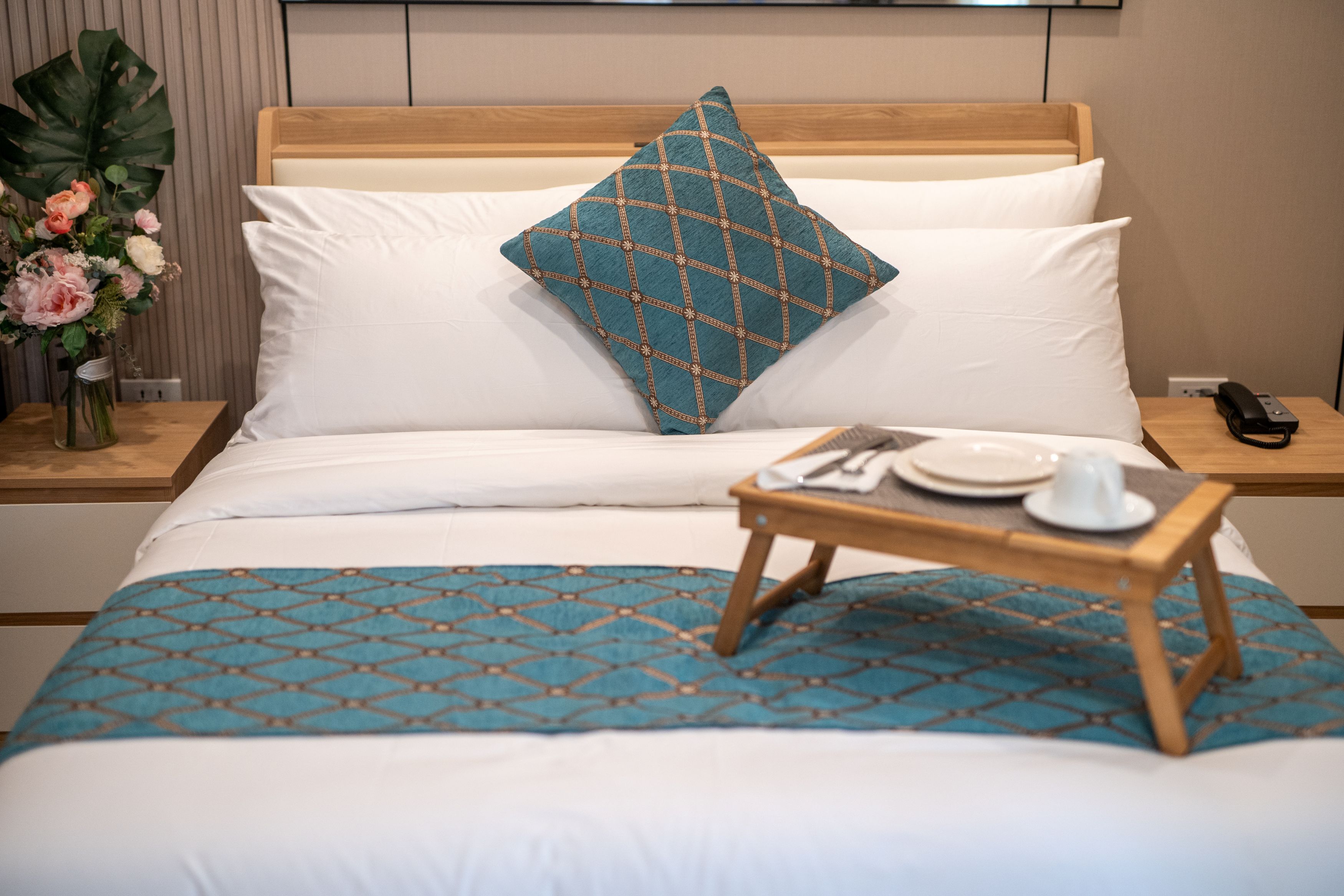 A neatly made bed with a patterned bedspread, a decorative pillow, and a bedside table with a tray of breakfast items.