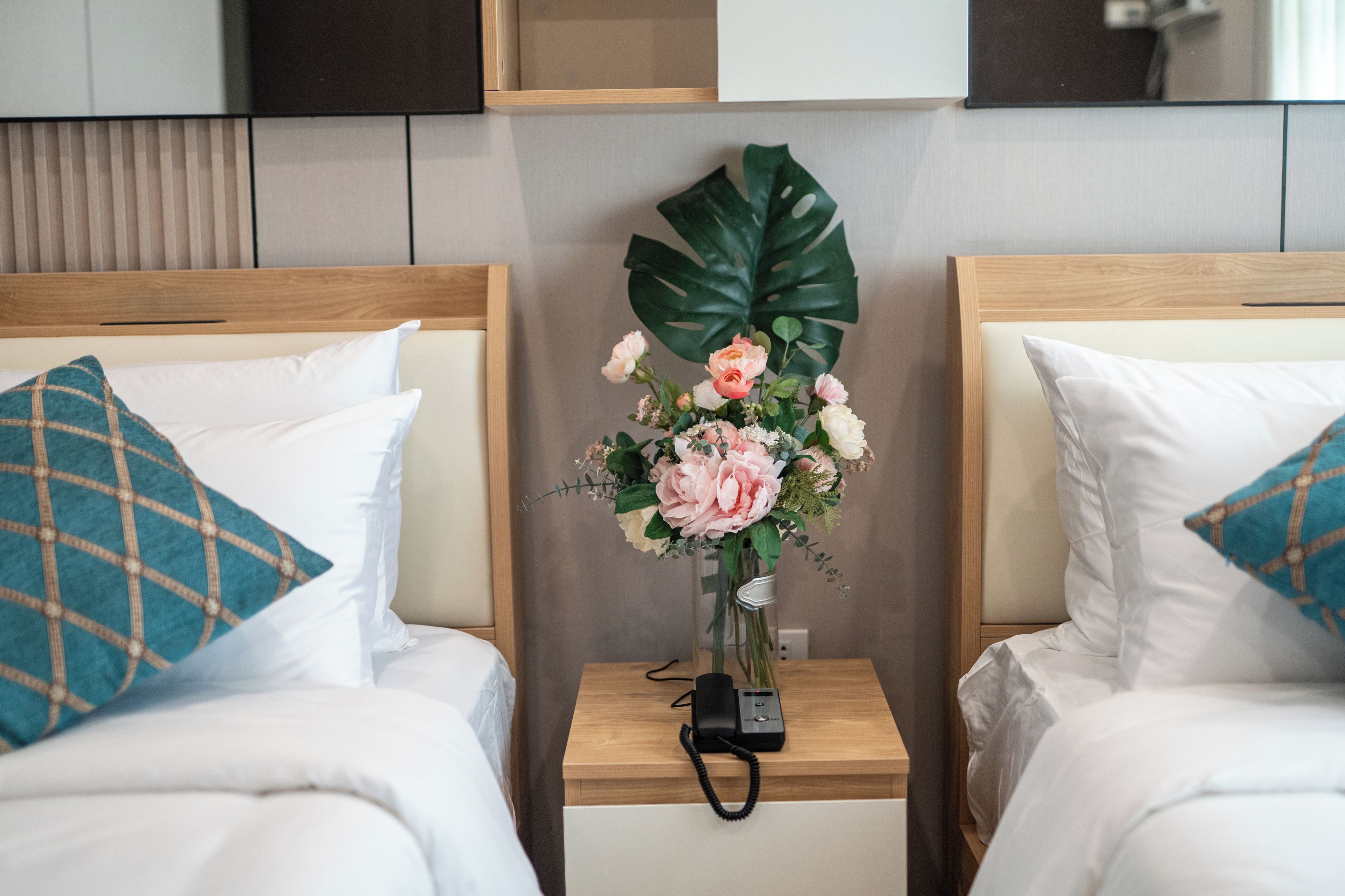 Una habitación de hotel con dos camas, cada una con una almohada y una mesita de noche. Un jarrón con flores está colocado sobre la mesa entre las camas.