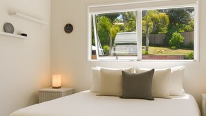 A bedroom with a large bed, a bedside table with a lamp, a window with a view of a backyard, and a wall-mounted shelf with decorative items. - La Maison Soleil with heated Swim Spa (Waiheke Island)