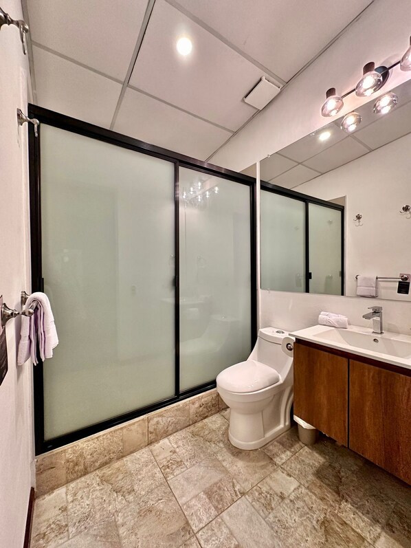 Un baño moderno con cabina de ducha de vidrio, inodoro, lavabo con tocador y un espejo grande.