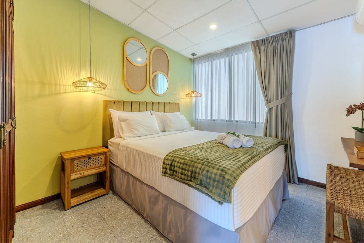 Une chambre d’hôtel avec un grand lit, deux lampes de chevet, un miroir, une fenêtre avec des rideaux et une petite table de chevet en bois.