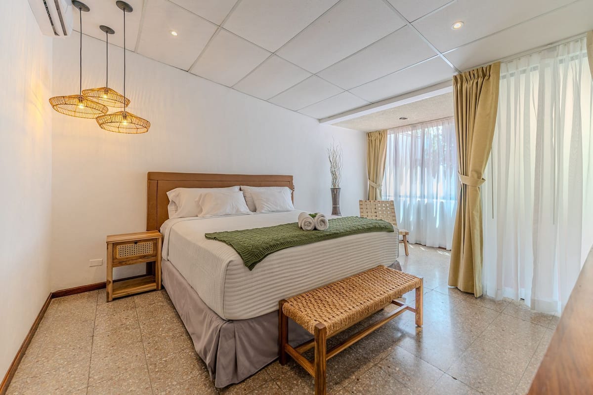 Une chambre d’hôtel avec un grand lit, une tête de lit en bois, des tables de chevet, un banc et une vue sur un balcon avec des rideaux.