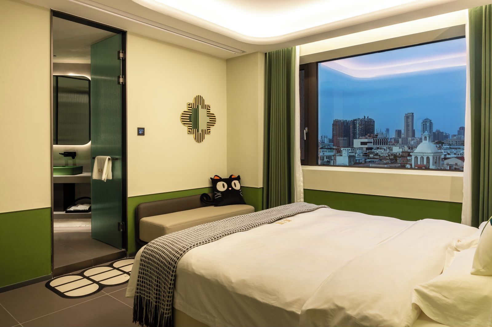A hotel room with a bed, a view of a cityscape, a green wall, and a black cat pillow.
