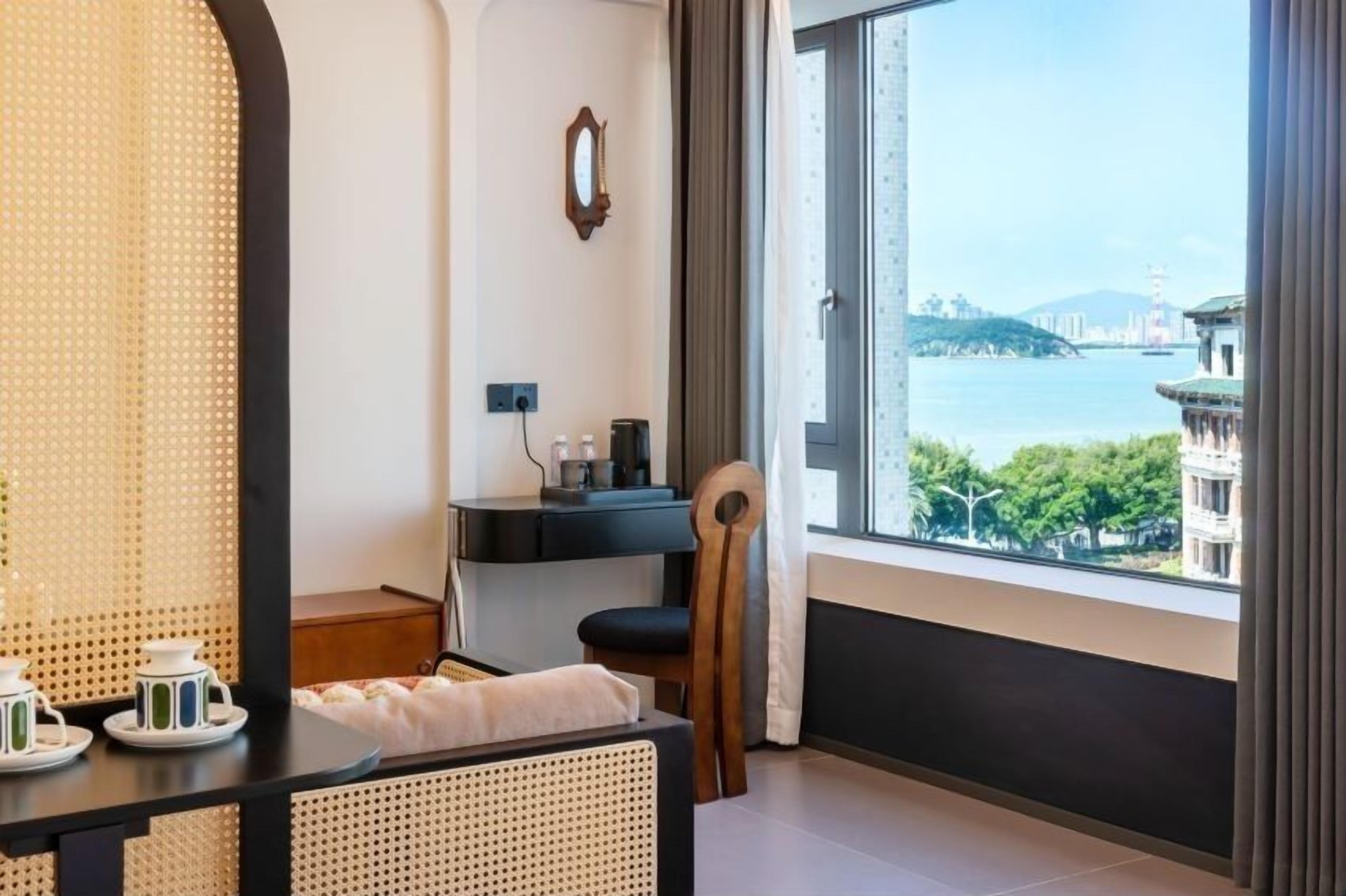 A hotel room with a large window offering a view of a body of water and buildings. There is a desk with a chair, a small table with teapots, and a bed with a woven headboard.