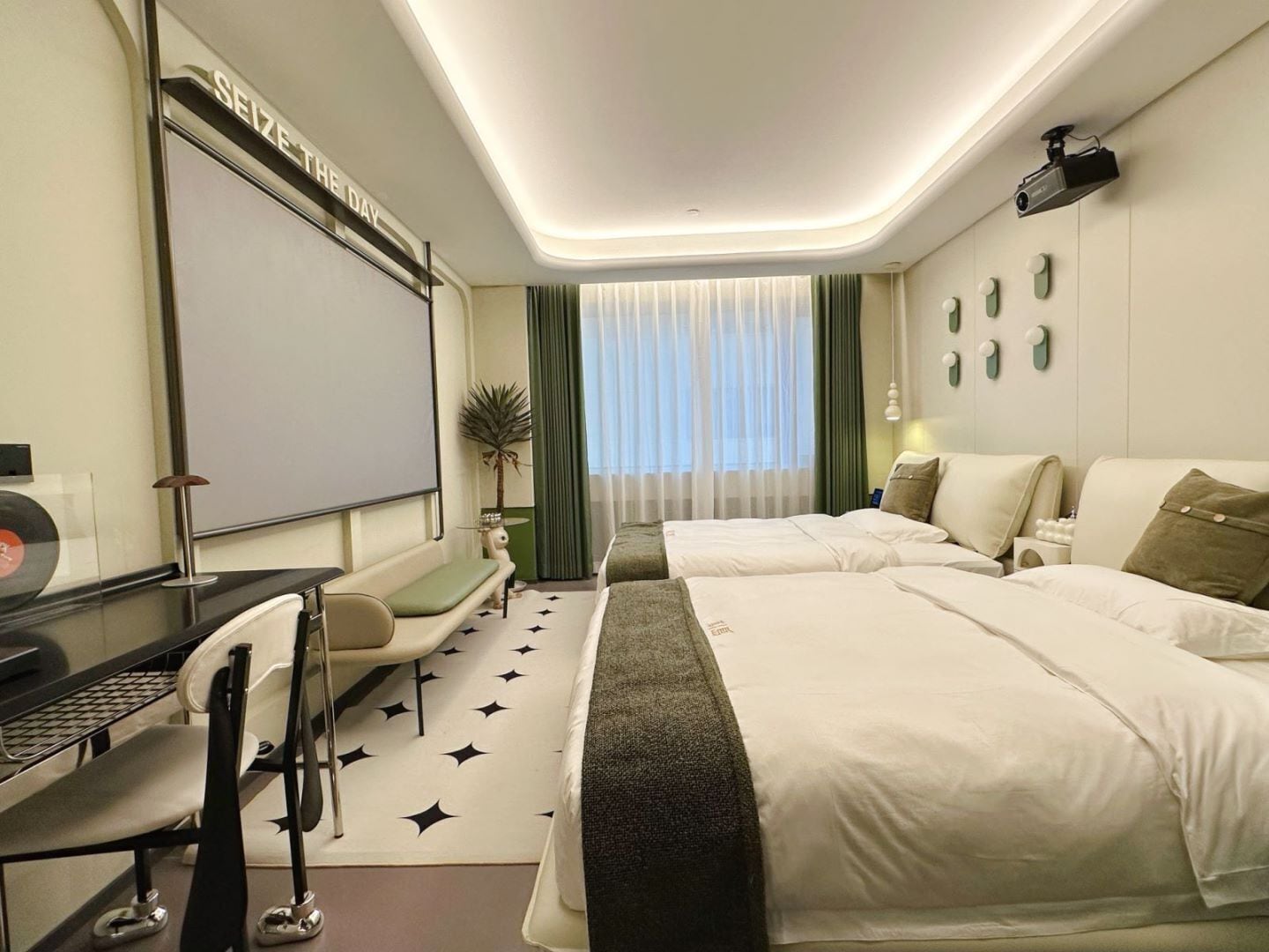 A hotel room with two beds, a desk with a computer, a projector screen, and a large window with curtains.