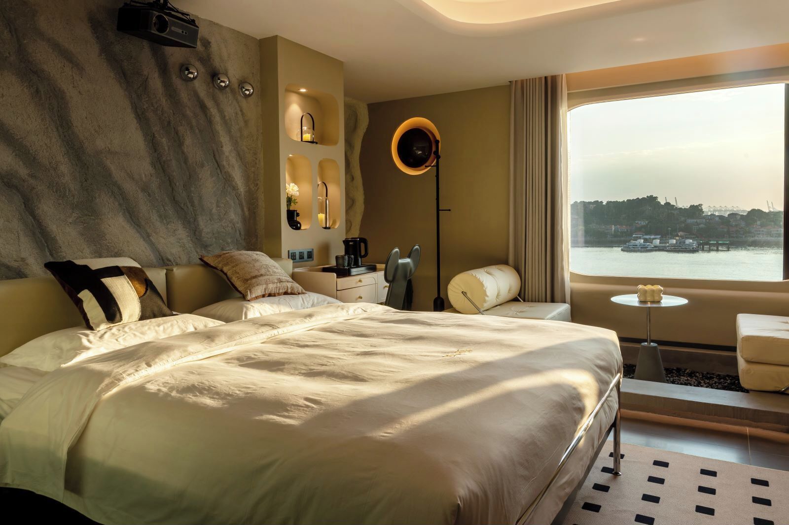 A modern hotel room with a large bed, a desk, a chair, a coffee maker, and a view of the city through the window.