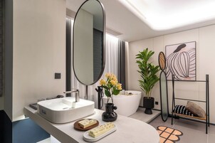 Un baño moderno con un espejo grande, lavabo blanco y una mesita de noche con una planta y un adorno.