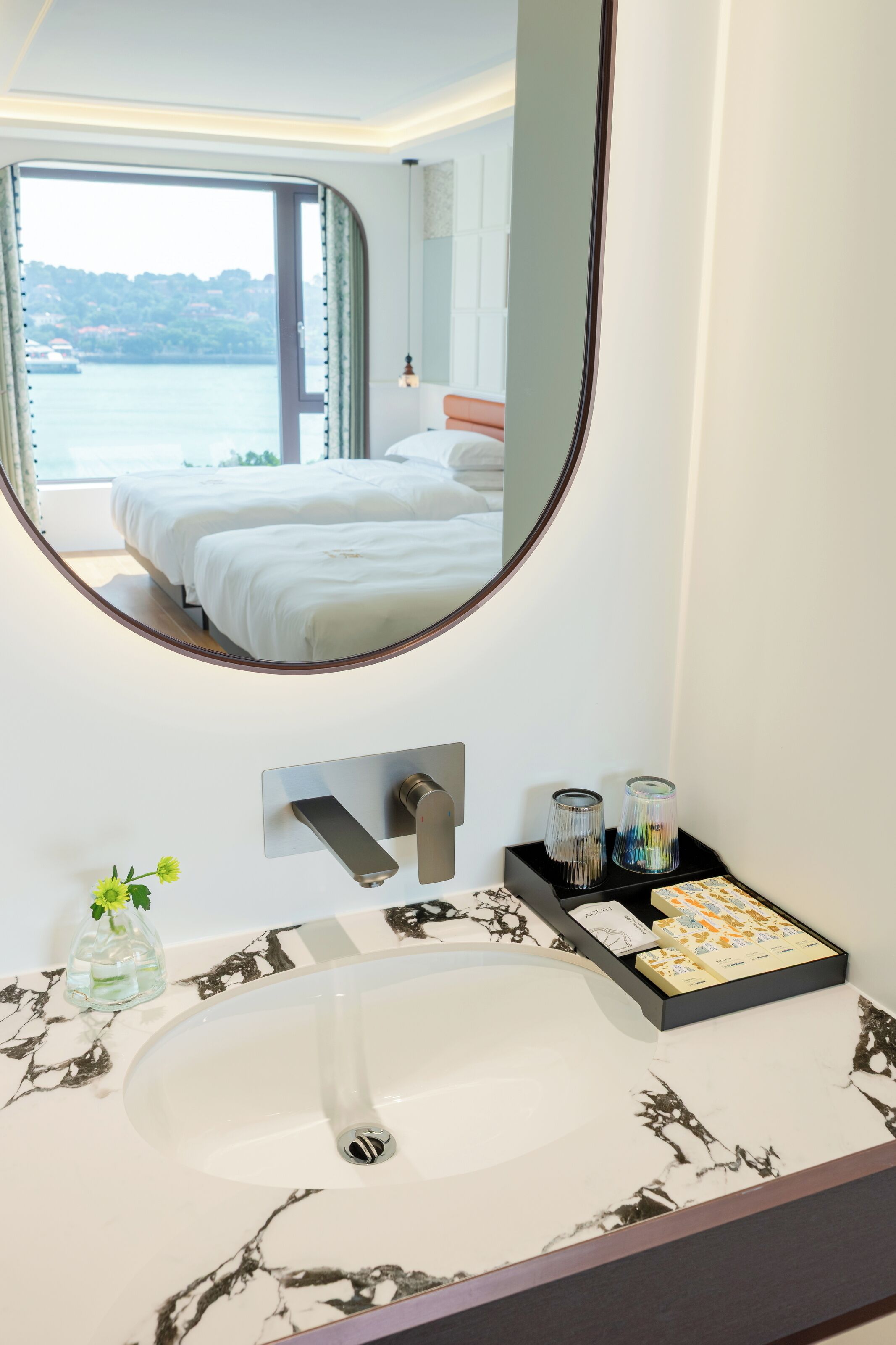 Une salle de bain d’hôtel dotée d’un grand miroir, d’un lavabo avec un plan de toilette en marbre et offrant une vue sur une chambre à coucher avec deux lits.