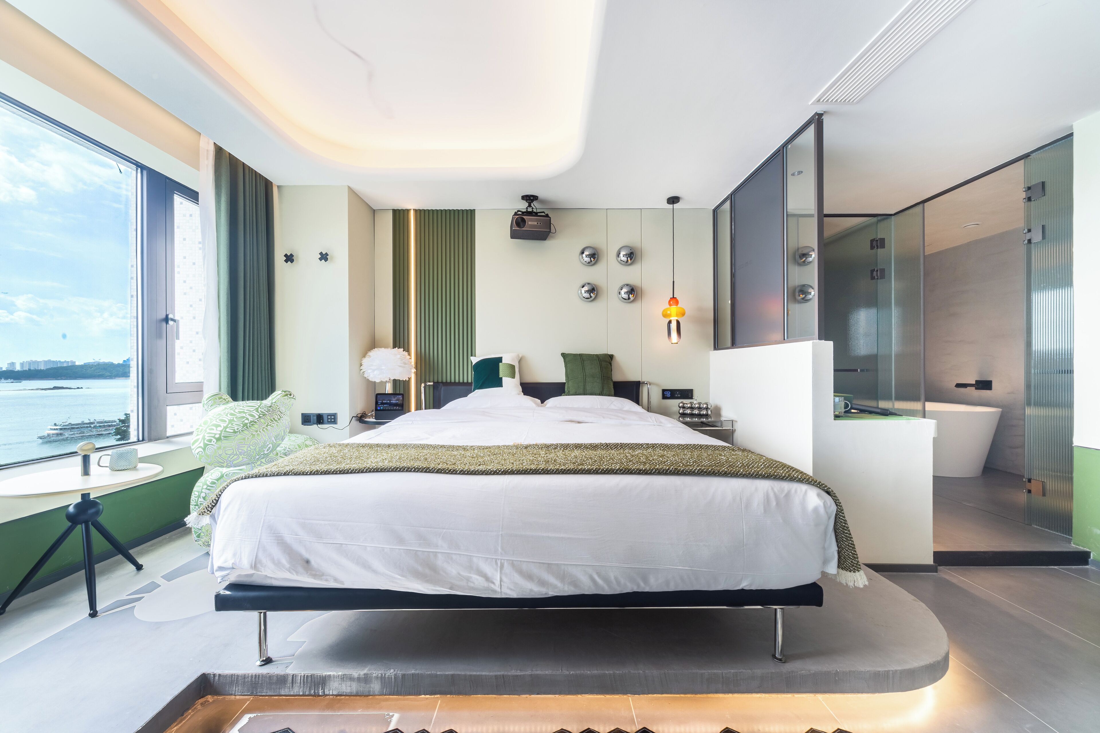 A modern hotel room with a large bed, a desk, a view of the water, and a glass-enclosed bathroom.
