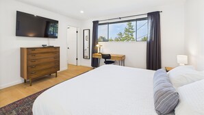 Un dormitorio con una cama grande, un aparador de madera, un escritorio con una silla, un televisor de pantalla plana en la pared y una ventana con cortinas.