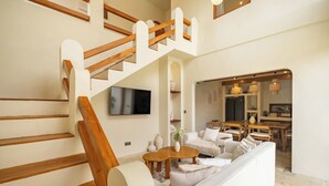 A modern living room with a staircase, a flat-screen TV, a sofa, a coffee table, and a dining area with a kitchen in the background. - 3BR Luxe Pool Villa Anaya (Canggu)
