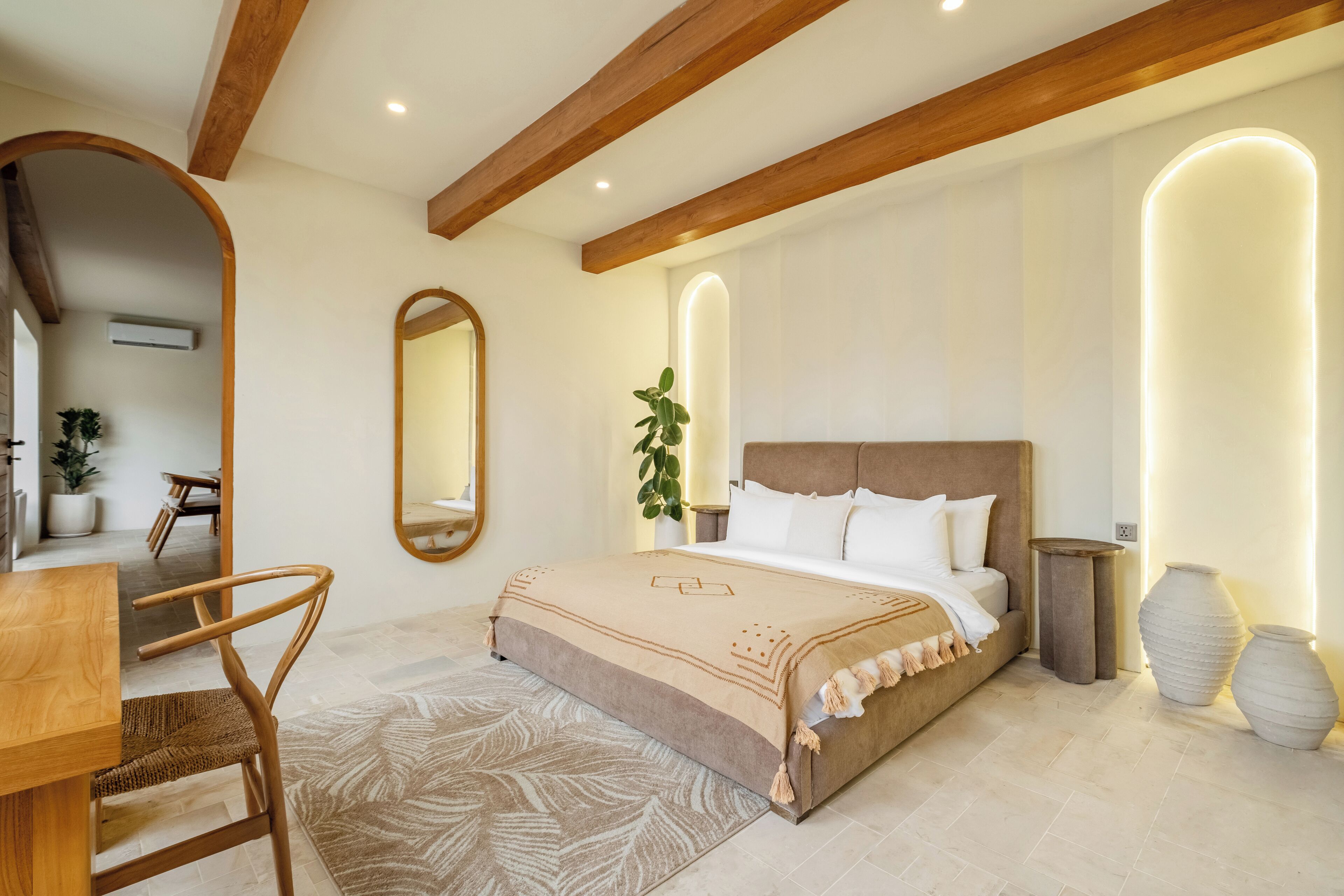 A bedroom with a bed, wooden ceiling beams, a mirror, a potted plant, and a bedside table.