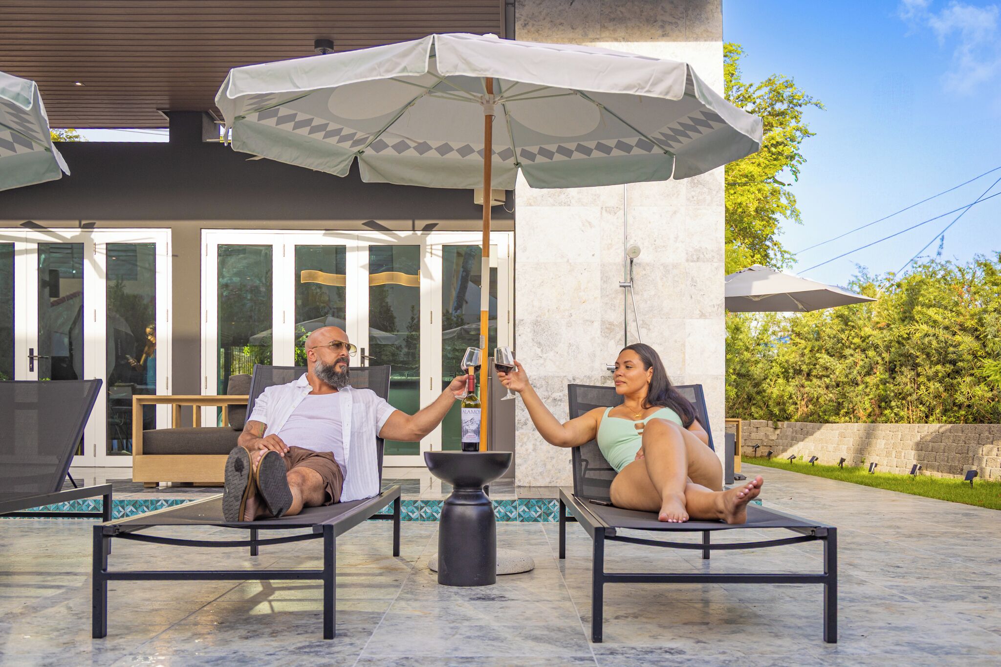 Deux personnes se détendent sur des chaises longues, à l’ombre d’un grand parasol. On aperçoit une piscine et un espace de détente extérieur en arrière-plan.