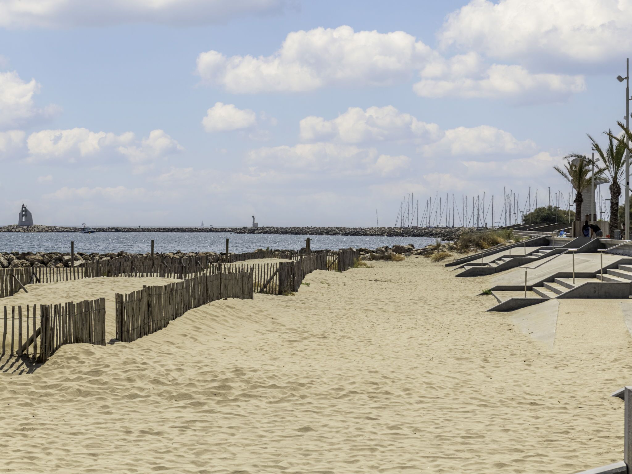 A sandy beach with a wooden fence, a lighthouse, and a rocky breakwater in the background.