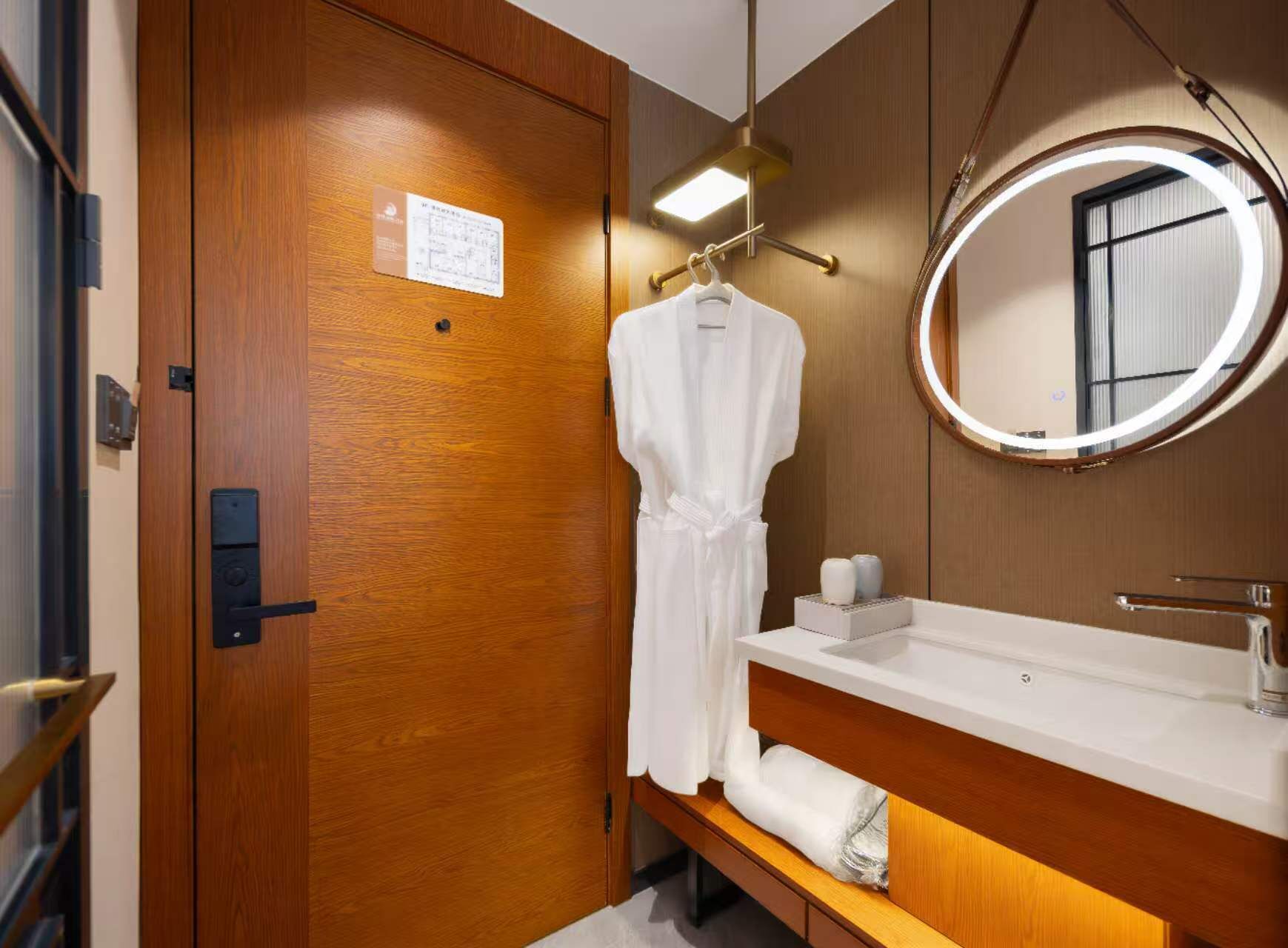 A hotel bathroom with a wooden door, a white robe hanging, a round mirror, and a sink with a towel rack.