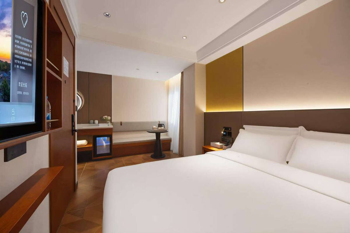 A hotel room with a large bed, a bedside table, a nightstand, a mirror, and a wall-mounted TV.