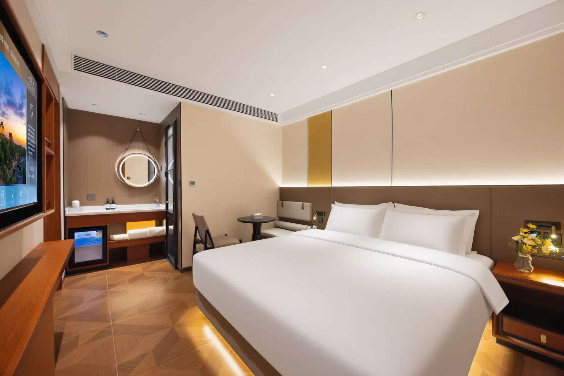A hotel room with a large bed, a bedside table with a lamp, a TV, a bathroom with a mirror, and a wooden floor.
