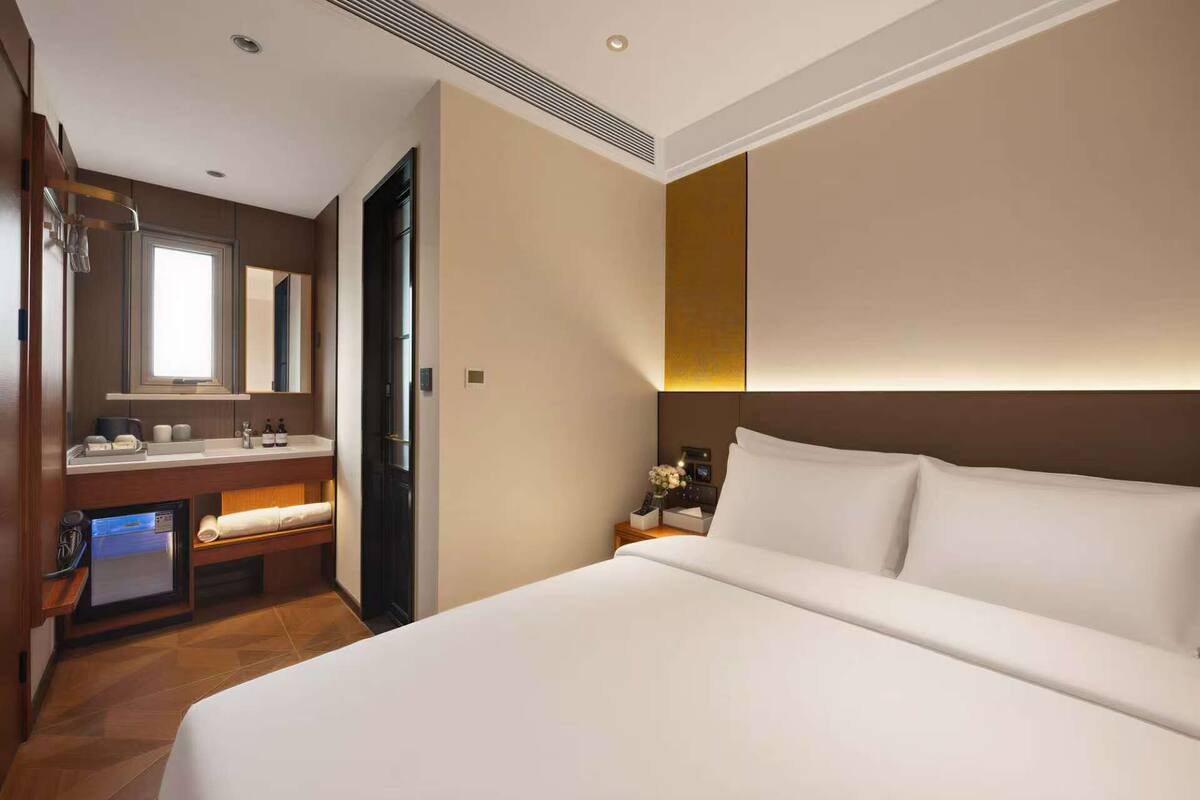 A hotel room with a bed, a vanity with a mirror, a small desk, and a wall-mounted light.