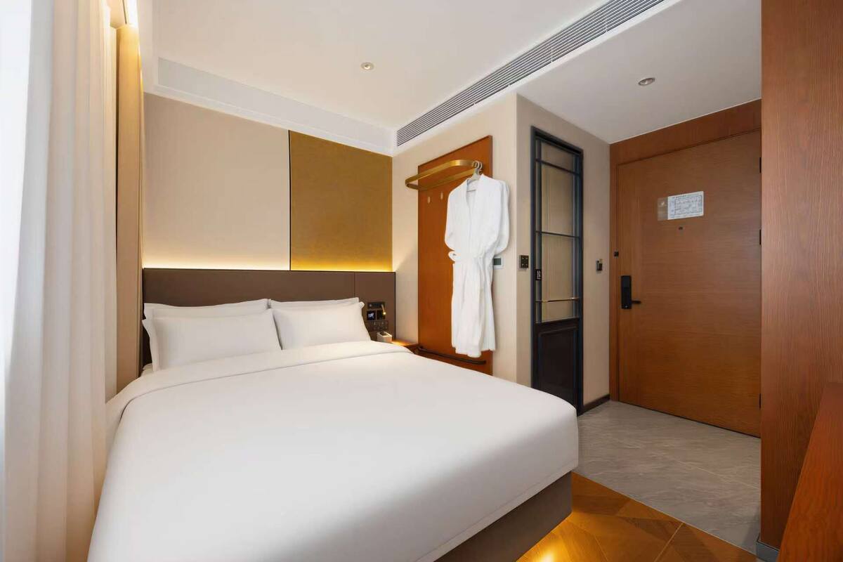 A hotel room with a bed, a robe hanging on a hook, a wooden door, and a wall-mounted light fixture.