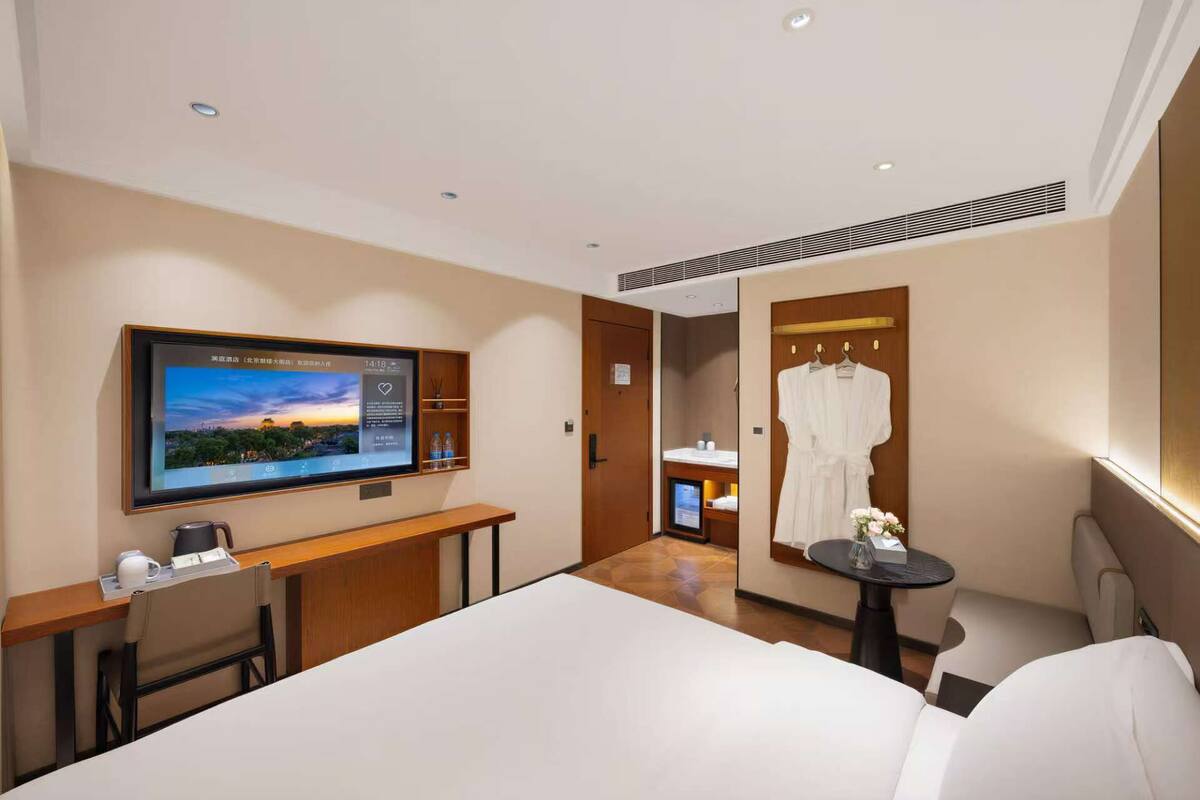 A hotel room with a bed, a TV mounted on the wall, a desk with a chair, a small table with a chair, and a robe hanging on the wall.
