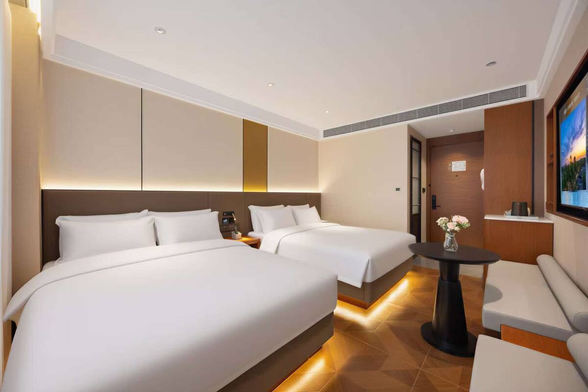 A hotel room with two beds, a small table with a vase, a sofa, and a TV on the wall.