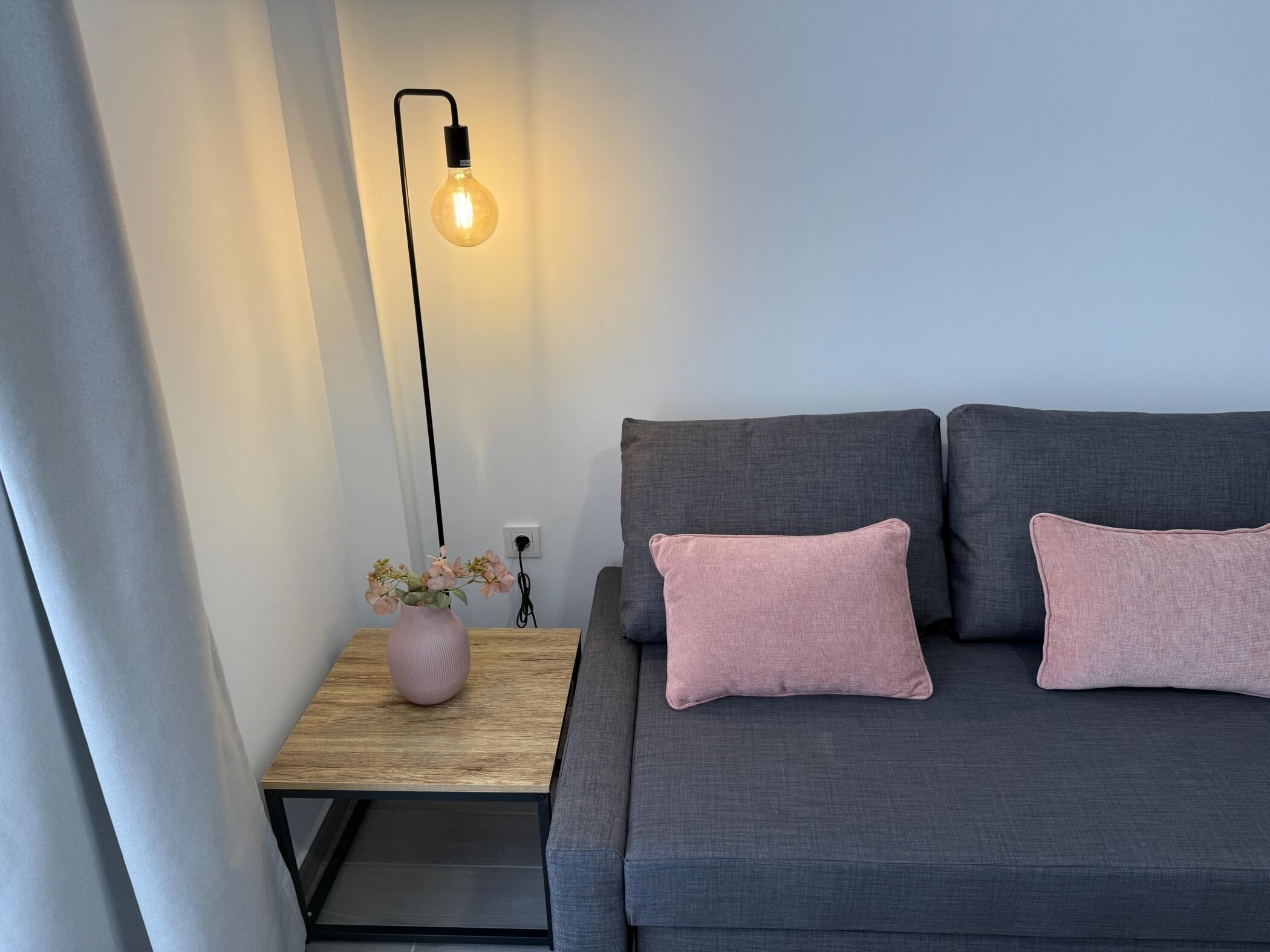 A modern living room with a grey sofa, two pink cushions, a wooden side table with a pink vase and flowers, and a floor lamp with a lit bulb.
