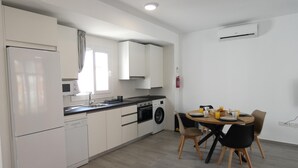 A compact kitchen with a refrigerator, stove, sink, and washing machine. There is a dining table with chairs and a fire extinguisher on the wall.