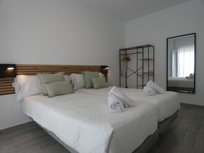 Una cama doble con cabecera de madera, ropa de cama blanca y almohadas verdes. Se observa una estantería metálica negra con un espejo y una toalla colgada.