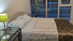 A bedroom with a bed, a glass table with a lamp, and a view of a balcony with blue railings.