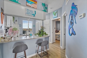 Una cocina con isla central, taburetes de bar, ventana con persianas y decoración en la pared.
