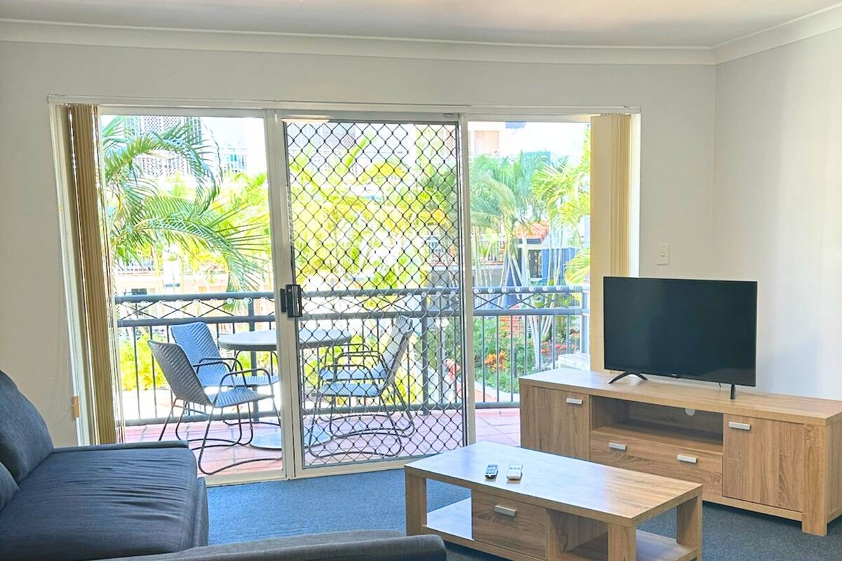 A room with a sofa, a coffee table, a TV on a wooden stand, and a sliding glass door leading to an outdoor balcony with chairs and plants.
