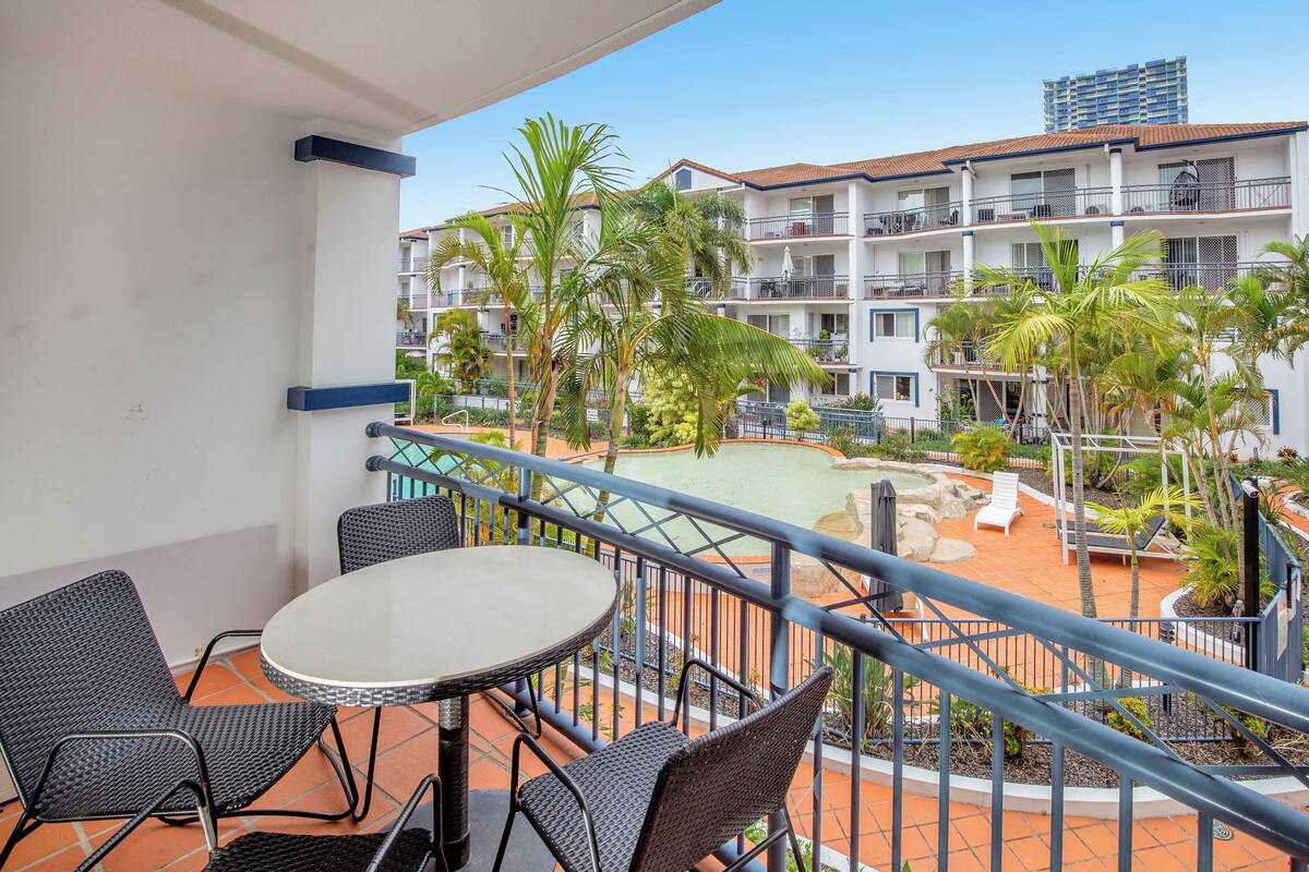 A balcony with a round table and chairs, overlooking a pool area with palm trees and a building in the background.