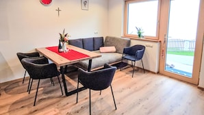 A modern living room with a wooden dining table, four chairs, a grey sofa, and a large window.