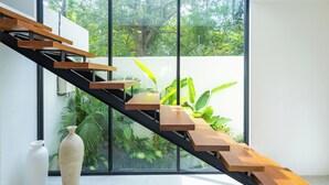 Una escalera moderna con escalones de madera y barandilla negra, que lleva a un balcón con vistas a vegetación exuberante.