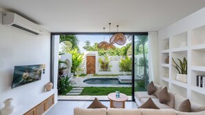 Un salón moderno con sofá, una mesa de centro y un televisor montado en la pared. Se ve un jardín con piscina a través de una ventana grande.