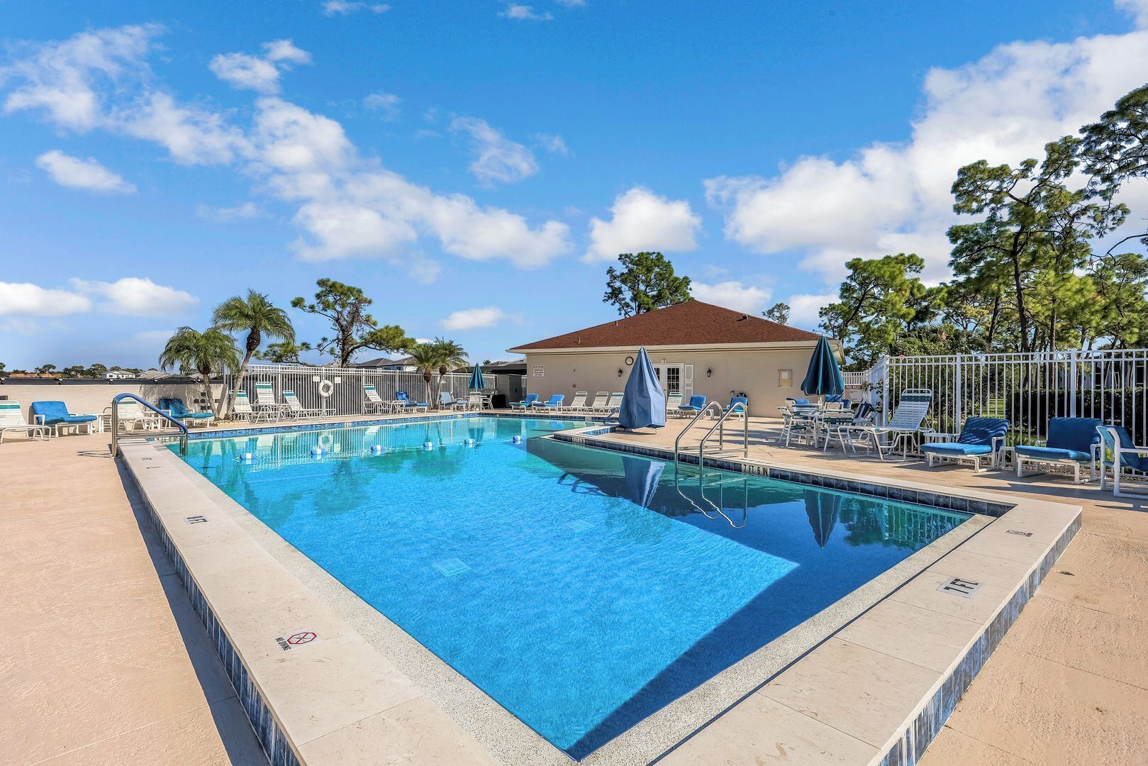 A rectangular swimming pool with a tiled edge, surrounded by lounge chairs and umbrellas, with a building in the background.